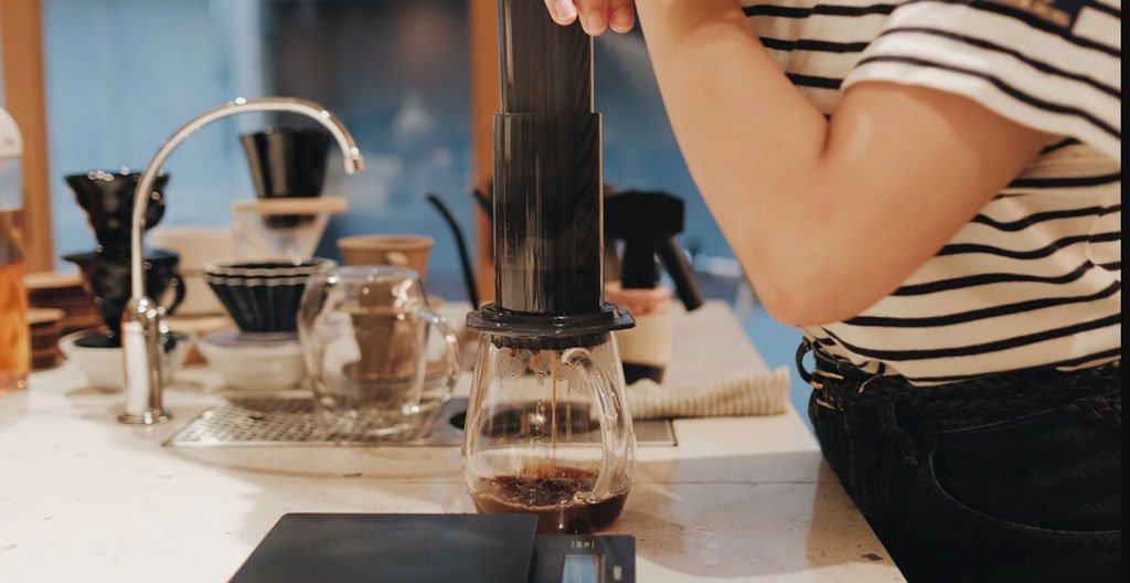 AeroPress en cours d'utilisation avec piston pressé, café s'écoulant dans une tasse, contexte nomade extérieur avec lumière matinale